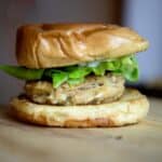 A close-up of a crab cake sandwich with a golden brown bun, leafy lettuce, sliced green pepper, and a thick, seasoned patty on a wooden surface.