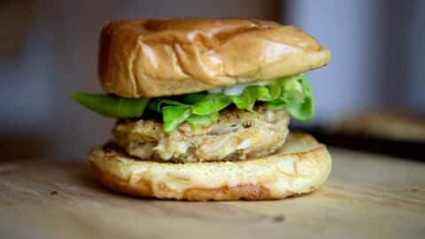 A close-up of a crab cake sandwich with a golden brown bun, leafy lettuce, sliced green pepper, and a thick, seasoned patty on a wooden surface.