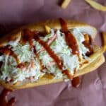 A hot dog topped with shredded coleslaw and drizzled with barbecue sauce, served in a bun with scattered French fries on brown paper—one of the 8 Super Bowl Food Ideas perfect for your game day spread.