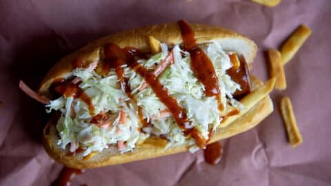 A hot dog topped with shredded coleslaw and drizzled with barbecue sauce, served in a bun with scattered French fries on brown paper—one of the 8 Super Bowl Food Ideas perfect for your game day spread.