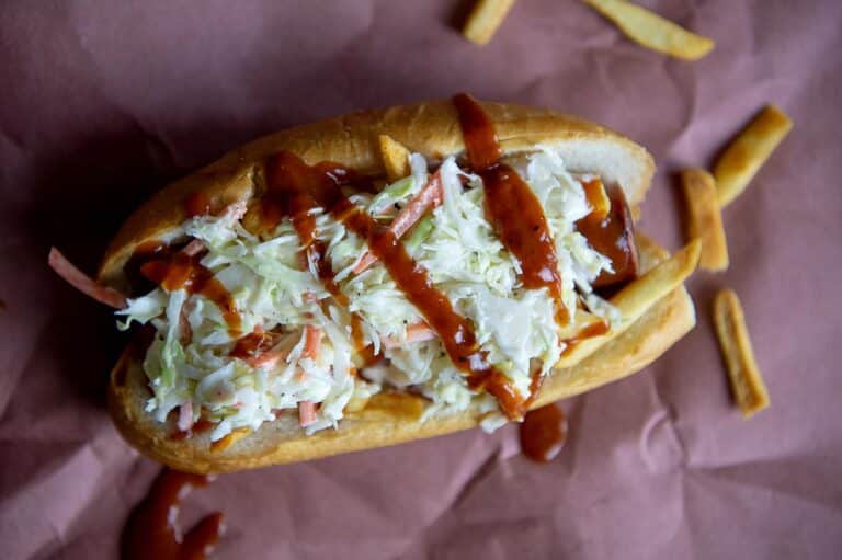 A hot dog inspired by the Polish Boy sandwich, topped with shredded coleslaw and drizzled with barbecue sauce, placed on brown paper, with a few French fries scattered around.