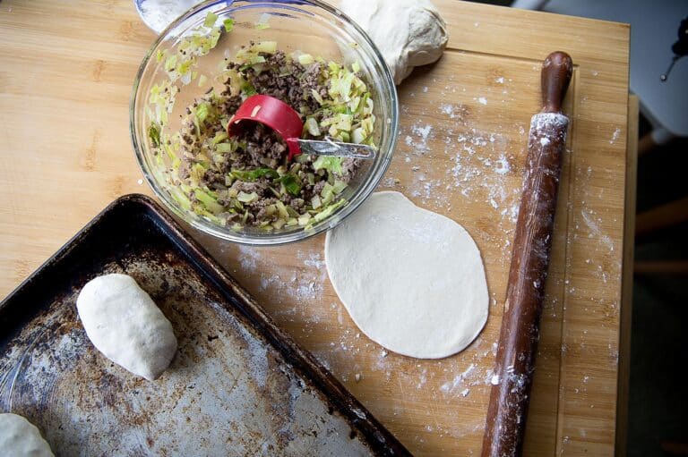 A wooden surface with rolled dough, a rolling pin, a bowl of chopped meat and vegetables, a red measuring cup, and a baking sheet with filled runza dough pieces, showing a delicious food preparation scene.