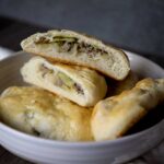 A bowl with three baked runza pastries, two of which are cut open to reveal a filling of ground meat and vegetables, likely cabbage or pickles. The pastries have a golden, slightly glossy crust and a soft, fluffy interior.