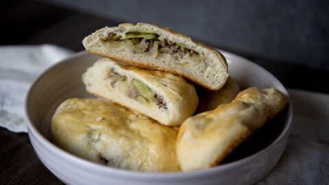 A bowl with three baked runza pastries, two of which are cut open to reveal a filling of ground meat and vegetables, likely cabbage or pickles. The pastries have a golden, slightly glossy crust and a soft, fluffy interior.