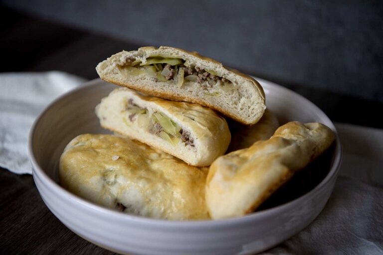 A bowl with three baked runza pastries, two of which are cut open to reveal a filling of ground meat and vegetables, likely cabbage or pickles. The pastries have a golden, slightly glossy crust and a soft, fluffy interior.