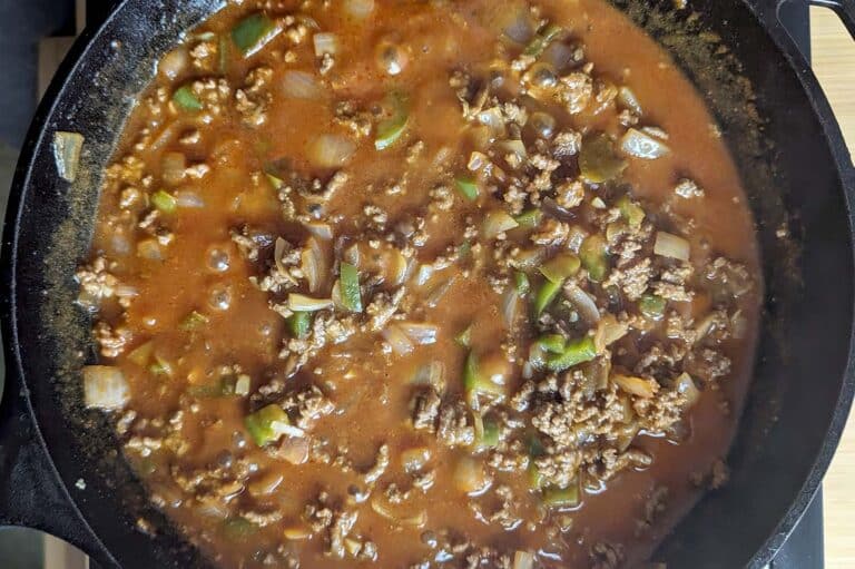 Ground beef, chopped onions, and green bell peppers simmer in a rich, tomato-based sauce in a black cast iron skillet for a classic sloppy joe flavor.