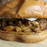 Close-up of a sloppy joe sandwich on a wooden surface, featuring a soft, golden bun filled with classic sloppy joe seasoned ground meat, diced onions, green peppers, and a layer of creamy mayonnaise.