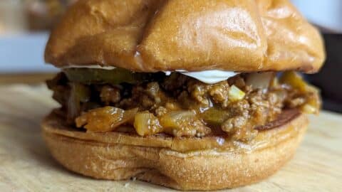Close-up of a sloppy joe sandwich on a wooden surface, featuring a soft, golden bun filled with classic sloppy joe seasoned ground meat, diced onions, green peppers, and a layer of creamy mayonnaise.