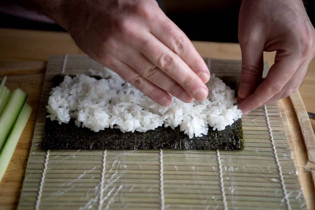 Hands of a sushi chef spreading sushi rice onto nori(seaweed). The nori is sitting on top of a saran wrapped bamboo mat.