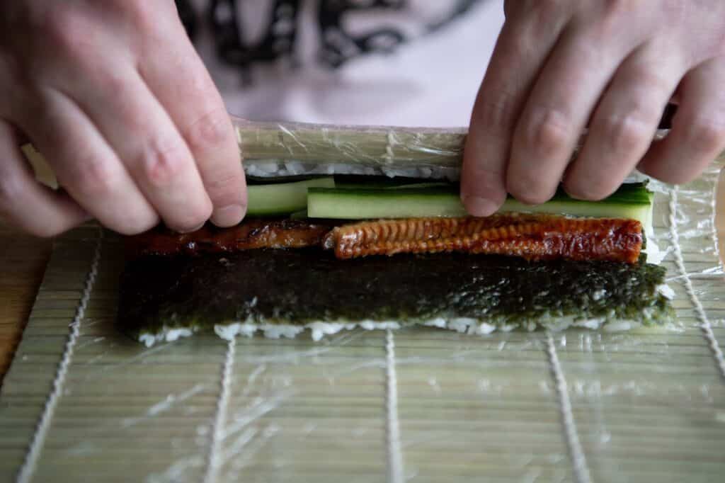 Nori is covered with sushi rice then flipped upside down while a chef places unagi and cucumber inside. They are preparing to make the sushi roll, otherwise known as maki.