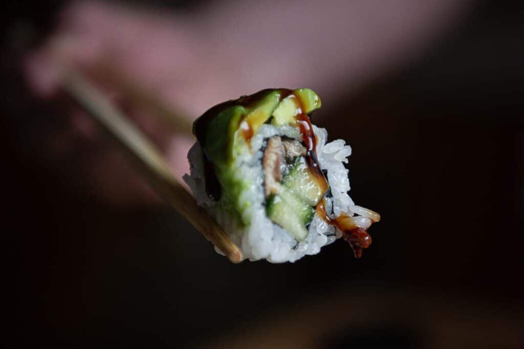 In this photo we can see a side shot of a cut piece of sushi. You can see the avocado resting on top of the roll, that has rice on the outside. On the inside you can see cucumber, and eel. You can also see a drizzle of eel sauce running down the face of the roll.