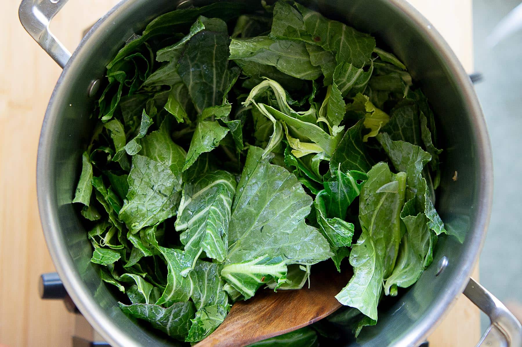 Collard greens in a huge pot being cooked down. You can also see a wooden spatula in the pot.