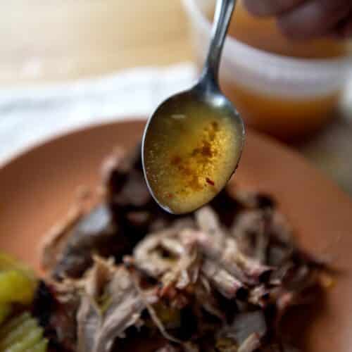 Smoked pork shoulder on a ceramic plate. On the side you can see pickle chips, and the chef pouring eastern north carolina bbq sauce over it with a spoon.