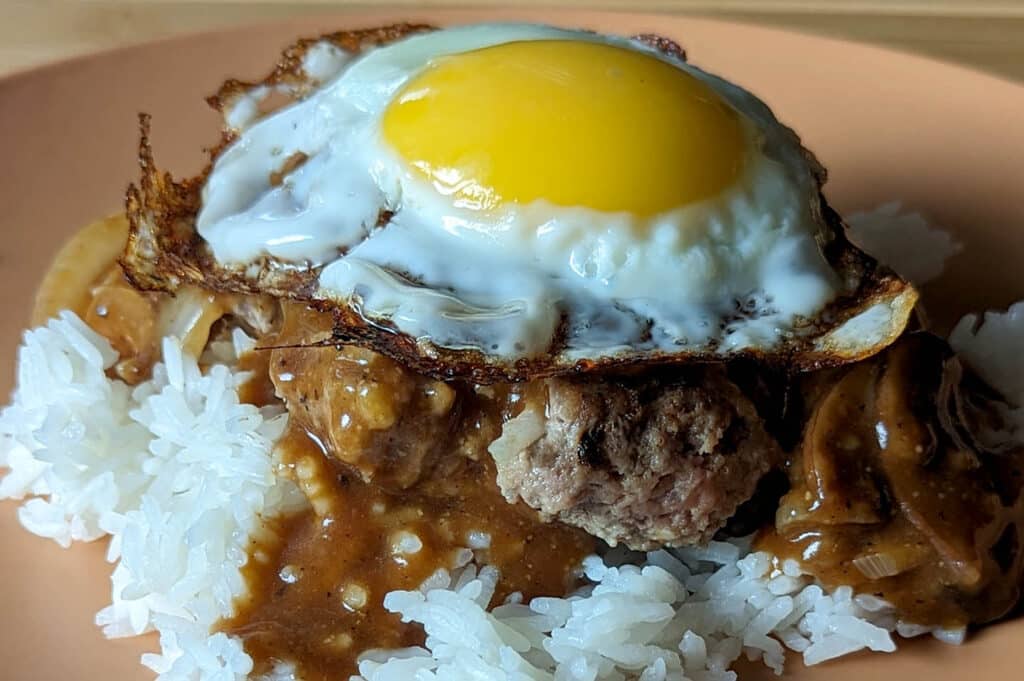 a sunny side up egg on top of a beef patty drenched in a brown gravy over white rice