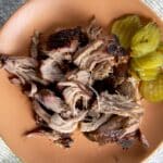 smoked pork butt on a ceramic plate with pickles on the side.