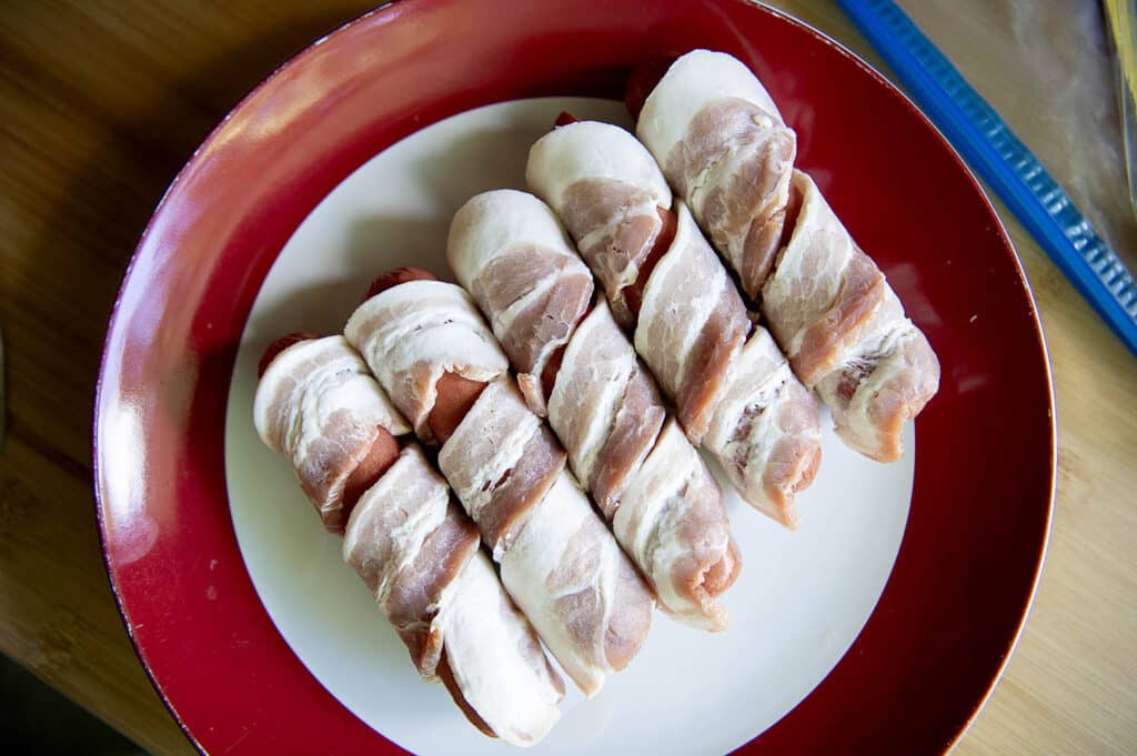 All beef hot dogs wrapped in bacon sitting on a plate, before being grilled.