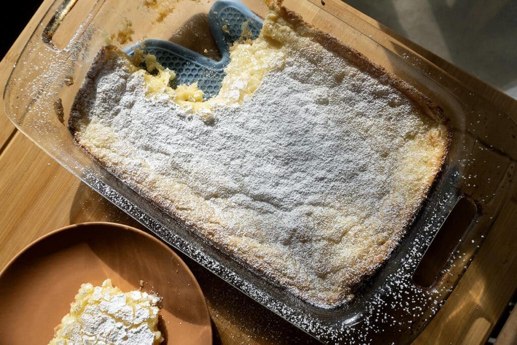 A glass baking dish filled with golden Ooey Gooey Butter Cake, topped with powdered sugar and missing a piece, sits on a wooden table. Nearby, a brown plate holds a single serving of this rich dessert.