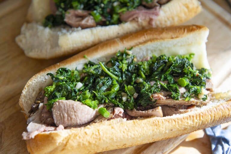 A close-up of a Roast Pork Sandwich or Italian sandwich with slices of roast beef and sautéed greens, served in a hoagie roll on a wooden surface.