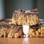 Three chocolate-topped Scotcheroos are stacked on a plate, with two at the bottom and one on top, showing their crunchy texture and glossy chocolate layer.