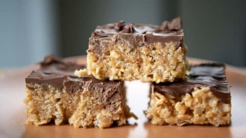 Three chocolate-topped Scotcheroos are stacked on a plate, with two at the bottom and one on top, showing their crunchy texture and glossy chocolate layer.