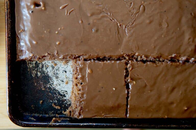 A baking sheet with a classic Texas Sheet Cake; several rectangular slices have been cut and removed, revealing the cake’s moist texture and rich, frosted surface.