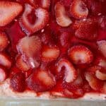 Sliced strawberries in a glossy red gelatin layer cover a creamy Strawberry Pretzel Salad base, viewed from above. The closely arranged strawberries create a vibrant and appetizing appearance.