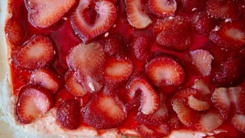 Sliced strawberries in a glossy red gelatin layer cover a creamy Strawberry Pretzel Salad base, viewed from above. The closely arranged strawberries create a vibrant and appetizing appearance.