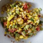 A white plate filled with a colorful mix of corn, tomatoes, okra, lima beans, yellow squash, onions, and bacon pieces, creating a vibrant and fresh summer succotash medley.