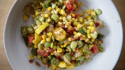 A white plate filled with a colorful mix of corn, tomatoes, okra, lima beans, yellow squash, onions, and bacon pieces, creating a vibrant and fresh summer succotash medley.