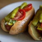 Two Chicago-Style Hot Dogs on a white plate, topped with sliced tomatoes, pickles, diced onions, relish, mustard, and nestled in classic poppy seed buns.