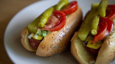 Two Chicago-Style Hot Dogs on a white plate, topped with sliced tomatoes, pickles, diced onions, relish, mustard, and nestled in classic poppy seed buns.