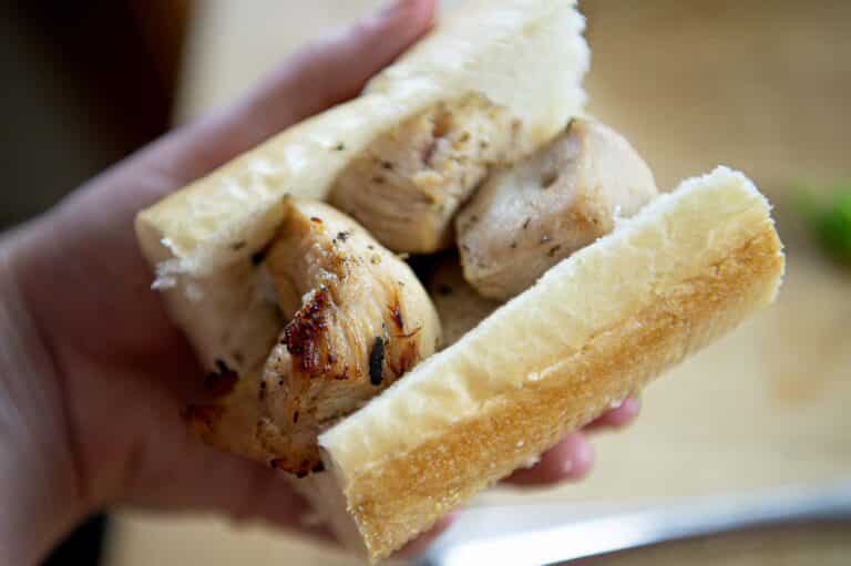 A hand holding a Chicken Spiedie sandwich, made with a thick baguette and chunks of grilled chicken. The sandwich is simple, with visible grill marks on the chicken and no additional fillings or condiments.