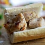 A close-up of a Chicken Spiedie sandwich filled with grilled chicken chunks inside a sliced baguette, resting on a wooden surface with blurred greens and food in the background.