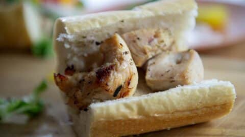 A close-up of a Chicken Spiedie sandwich filled with grilled chicken chunks inside a sliced baguette, resting on a wooden surface with blurred greens and food in the background.