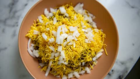 A bowl filled with shredded yellow cheese and chopped white onions, perfect toppings for Cincinnati Chili, sits on top of a marble surface.