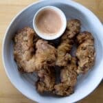 A white bowl with five pieces of fried chicken, including crispy Finger Steaks, and a small cup of pink dipping sauce, placed on a light wooden surface.