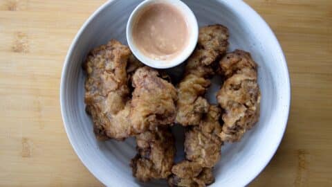 A white bowl with five pieces of fried chicken, including crispy Finger Steaks, and a small cup of pink dipping sauce, placed on a light wooden surface.