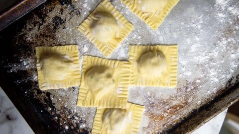 Six uncooked square homemade ravioli are arranged in a cross shape on a floured metal baking sheet, ready to be cooked.
