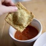 A hand holds a piece of breaded, toasted ravioli air fryer style above a bowl of marinara sauce on a wooden table.