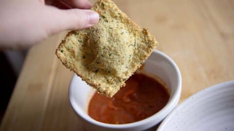 A hand holds a piece of breaded, toasted ravioli air fryer style above a bowl of marinara sauce on a wooden table.