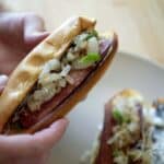 A person holds a Seattle-Style Hot Dog in a bun topped with chopped grilled onions and green peppers. Another hot dog with similar toppings sits on a plate in the background.