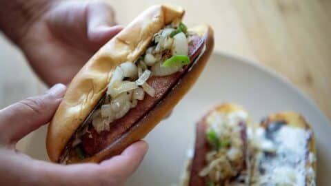 A person holds a Seattle-Style Hot Dog in a bun topped with chopped grilled onions and green peppers. Another hot dog with similar toppings sits on a plate in the background.