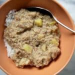 A bowl of white rice topped with thick, brownish South Carolina BBQ Hash containing chunks of vegetables, served with a metal spoon in an orange bowl on a marble surface.