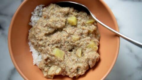A bowl of white rice topped with thick, brownish South Carolina BBQ Hash containing chunks of vegetables, served with a metal spoon in an orange bowl on a marble surface.
