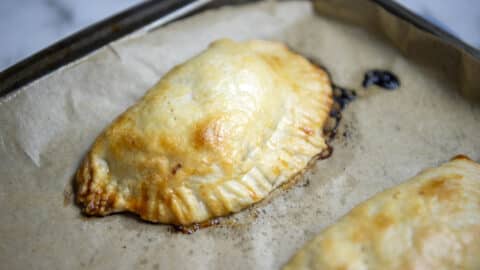 A golden-brown baked hand pie, inspired by a classic pasty recipe, rests on parchment paper atop a baking sheet, its slightly crimped edges framing a small amount of filling oozing out.