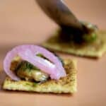 A close-up of a cracker topped with smoked oysters, pickled red onion, herbs, and a savory spread on a peach-colored plate, with a blurred hand preparing another similar cracker in the background.