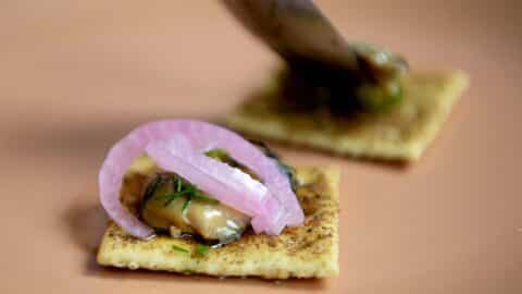 A close-up of a cracker topped with smoked oysters, pickled red onion, herbs, and a savory spread on a peach-colored plate, with a blurred hand preparing another similar cracker in the background.