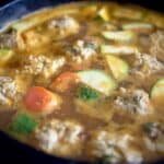 A close-up of a pot filled with a simmering soup containing meatballs, sliced zucchini, and carrots in a rich, golden broth.