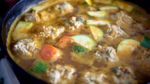 A close-up of a pot filled with a simmering soup containing meatballs, sliced zucchini, and carrots in a rich, golden broth.