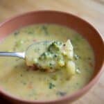 A spoonful of creamy broccoli cheddar soup is held above a bowl, showing chunks of broccoli and carrots in a thick yellow broth—an irresistible Broccoli Cheddar Soup Recipe come to life.